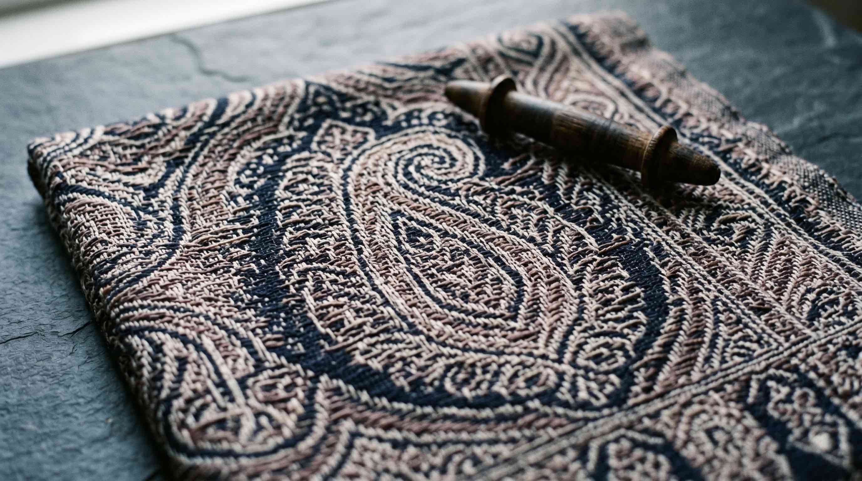Close-up of a Kani-woven pashmina shawl in midnight navy with complex paisley motifs in warm brown tones, with a small wooden kani spool resting on the surface — showing the woven-in pattern that defines authentic Kani work.