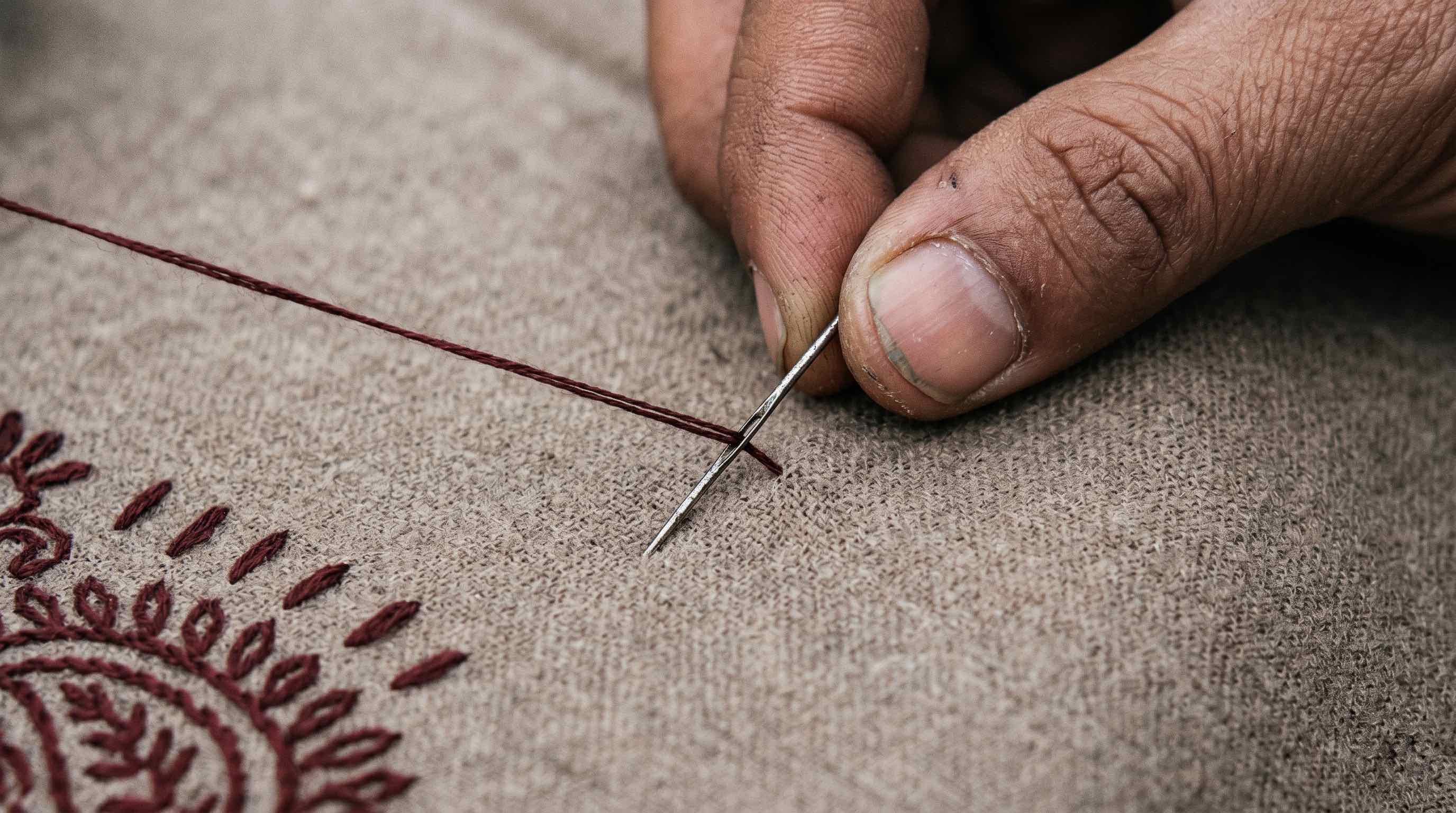A karigar's hand mid-stitch — a sozni needle entering pale taupe pashmina cloth with dark claret thread pulled taut, with partial floral border embroidery visible in the lower left corner.